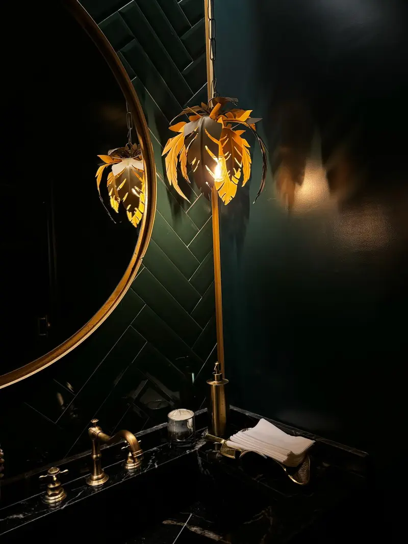 Toilette con pared verde oscuro, espejo redondo de marco dorado y apliques dorados con forma de hojas.